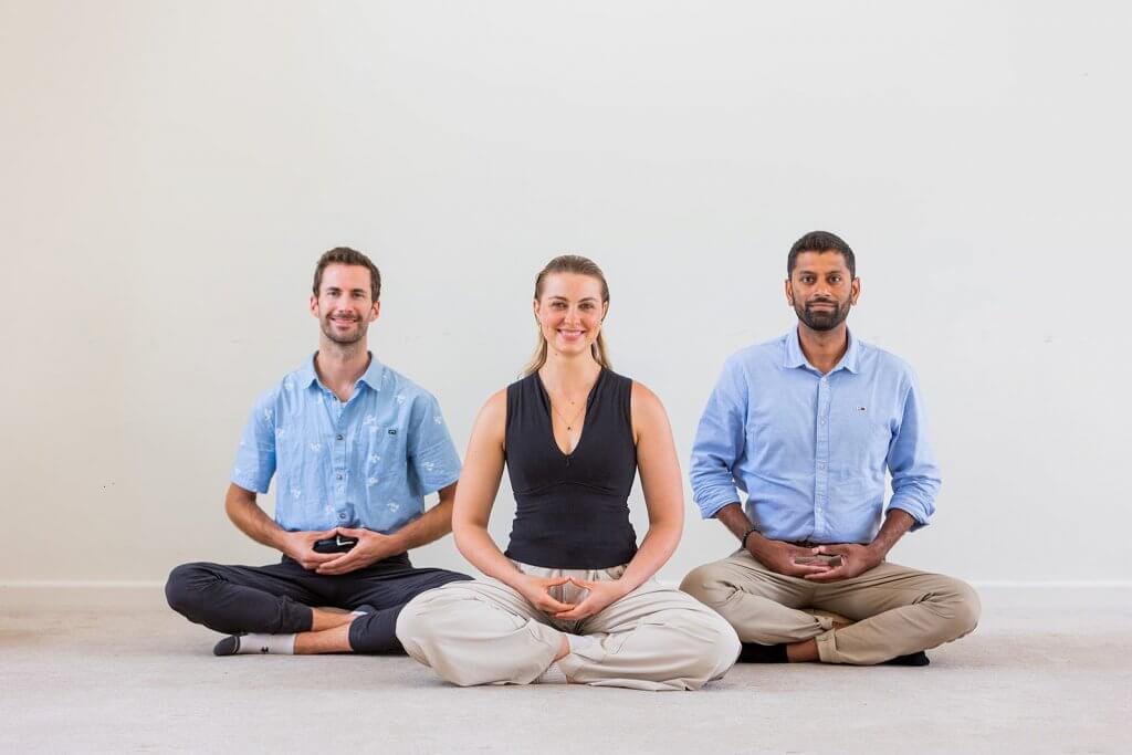 Three people sat facing the camera smiling and sitting in a meditation posture