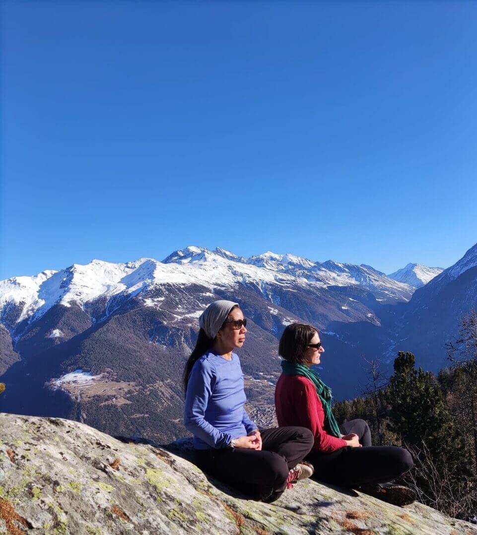 Two people peacefully meditating with their eyes closed on a mountainside with a clear blue sky in the background.