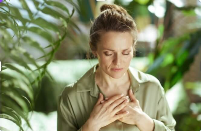 Woman looking serene and peaceful surrounded by greenery, sh has eyes closed and her hands at her heart