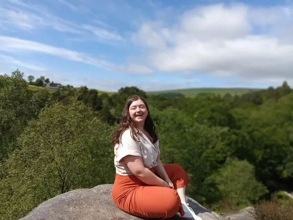 a smiling young lady sat cross legged on top of a rock with a background of trees and blue sky.