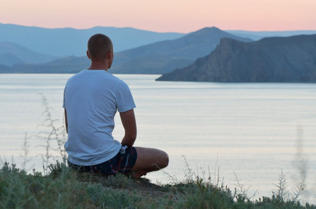 A man meditating peacefully overlooking a lake with mountains in the background.